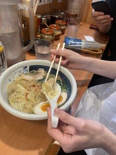 two hands holding chopsticks over a bowl of noodles with an egg in it