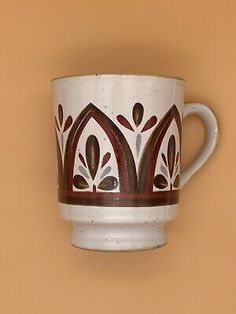 a white coffee cup with brown designs on the rim and bottom is hanging from a wall