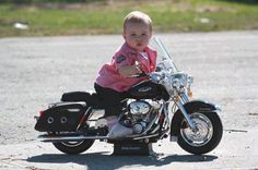 a baby sitting on top of a motorcycle in the middle of a parking lot with grass behind it