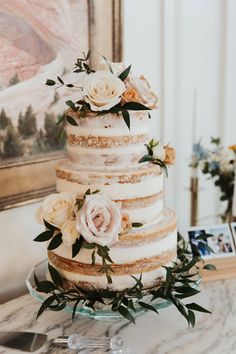 a three tiered cake with flowers and greenery sits on a table in front of a painting