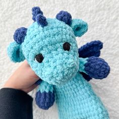 a hand holding a blue crocheted dragon stuffed animal with black eyes and tail