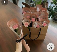 a hand holding a bouquet of pink roses on top of a wooden table next to a brown paper bag