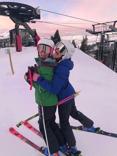 two people hugging each other on skis at the top of a slope with a chair lift in the background