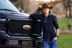 a man wearing a cowboy hat leaning on the hood of a black pick up truck