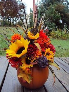 a pumpkin filled with lots of flowers sitting on top of a wooden table next to grass