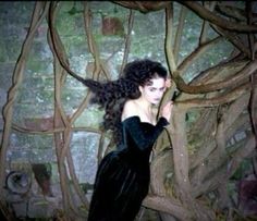 a woman with long hair standing in front of a wall covered in vines and branches