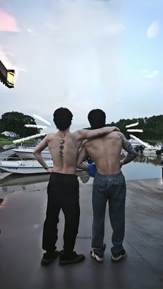 two young men standing next to each other in front of a body of water with boats behind them