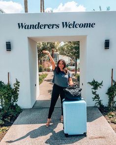 a woman is standing in front of a welcome sign with her hand up and holding a suitcase