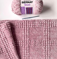 a pink crochet blanket next to a ball of yarn and knitting needles on a white surface