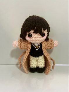 a small crocheted doll is posed on a white surface, wearing a brown jacket and black shirt