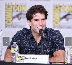 two men sitting at a table with water bottles in front of them and one man holding a microphone