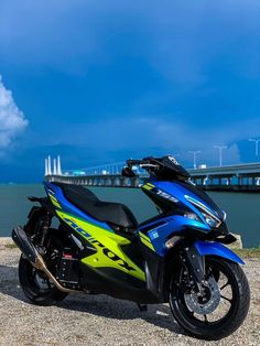 a blue and yellow motorcycle parked next to the ocean with a bridge in the background