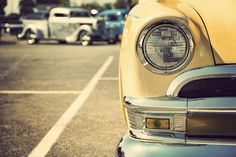 the front end of an old yellow car parked in a parking lot with other cars