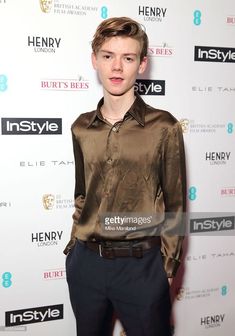 a young man in a brown shirt and black pants poses on the red carpet at an event