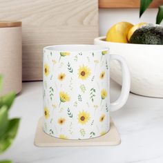 a white coffee mug sitting on top of a counter next to a bowl of fruit