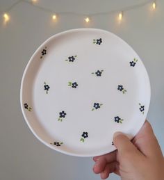 a hand holding a white plate with black and green flowers painted on it in front of a string of lights