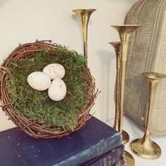 three eggs in a nest on top of a book next to two candlesticks