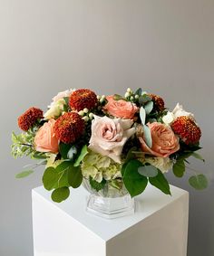 a vase filled with lots of flowers on top of a white cubed table next to a gray wall