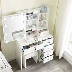 a white vanity table with drawers and lights on the top, in front of a window