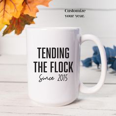 a white coffee mug with the words tending the flock since 2013 on it next to fall leaves