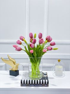 pink tulips are in a clear vase on a white table next to a gold bird