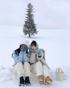 two people sitting in the snow next to a small tree with one person holding a stuffed animal