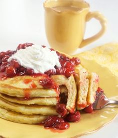 a stack of pancakes topped with whipped cream and cherries on a yellow plate next to a cup of coffee