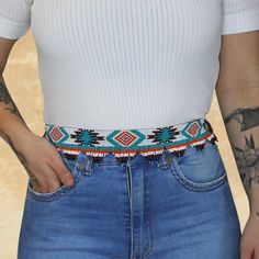 a woman wearing a white shirt and blue jeans with tattoos on her arm, holding onto a colorful beaded belt