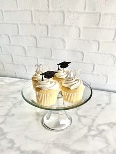 two cupcakes with white frosting and black graduation caps on them sitting on a glass plate