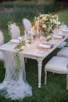 the table is set with white chairs and candles on it, along with greenery