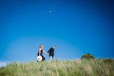 a man and woman holding hands walking through tall grass on a hill with a moon in the background