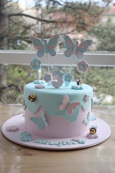 a blue and pink cake with butterflies on top is sitting on a table next to a window