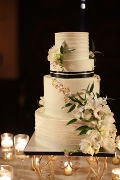 a wedding cake with white flowers and greenery on the top is surrounded by candles