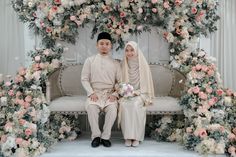 a man and woman sitting on a bench in front of a flower covered arch with flowers