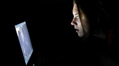 a man sitting in the dark looking at his computer screen and holding it up to his face