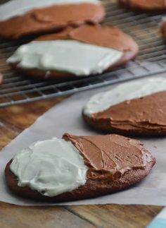 chocolate cookies with white frosting sitting on top of a wooden table next to a cooling rack