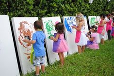 several children are painting on the side of white boards in front of a hedge wall