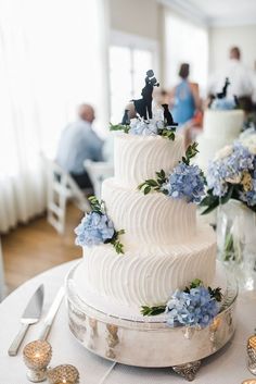 a white wedding cake with blue flowers and figurines on top is sitting on a table