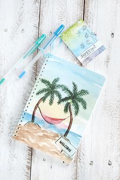 an open notebook with palm trees on the beach and watercolor pencils next to it