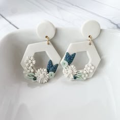 two white hexagonal earrings with flowers and leaves hanging from them on a plate