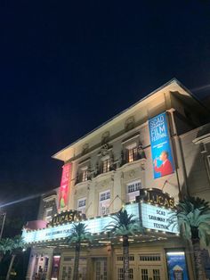 the front of a large building at night with palm trees and lights on it's sides