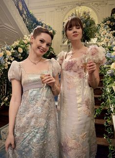 two women in dresses standing next to each other holding drinks and posing for the camera