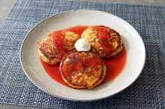 three pancakes on a white plate with red sauce and whipped cream in the middle, sitting on a blue place mat