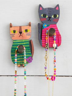 two colorful cat wall hangings on the side of a white wooden wall with beads and chains attached to them