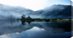 a lake surrounded by mountains and fog in the air with trees on both sides that are reflected in the water