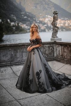 a woman in a black and grey dress standing on a balcony near the water with mountains behind her