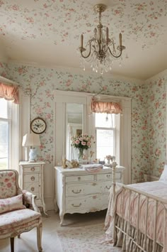 a bedroom with floral wallpaper and pink bedding, chandelier, chair, dresser and mirror