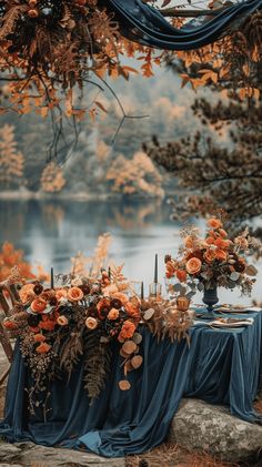 an outdoor table set up with blue cloths and orange flowers on it, next to a lake