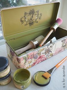 an open box with paint, brushes and other items in it sitting on a table next to a window