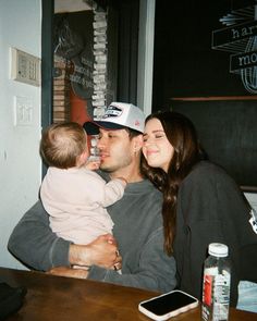 a man holding a baby while sitting next to a woman at a table with a cell phone